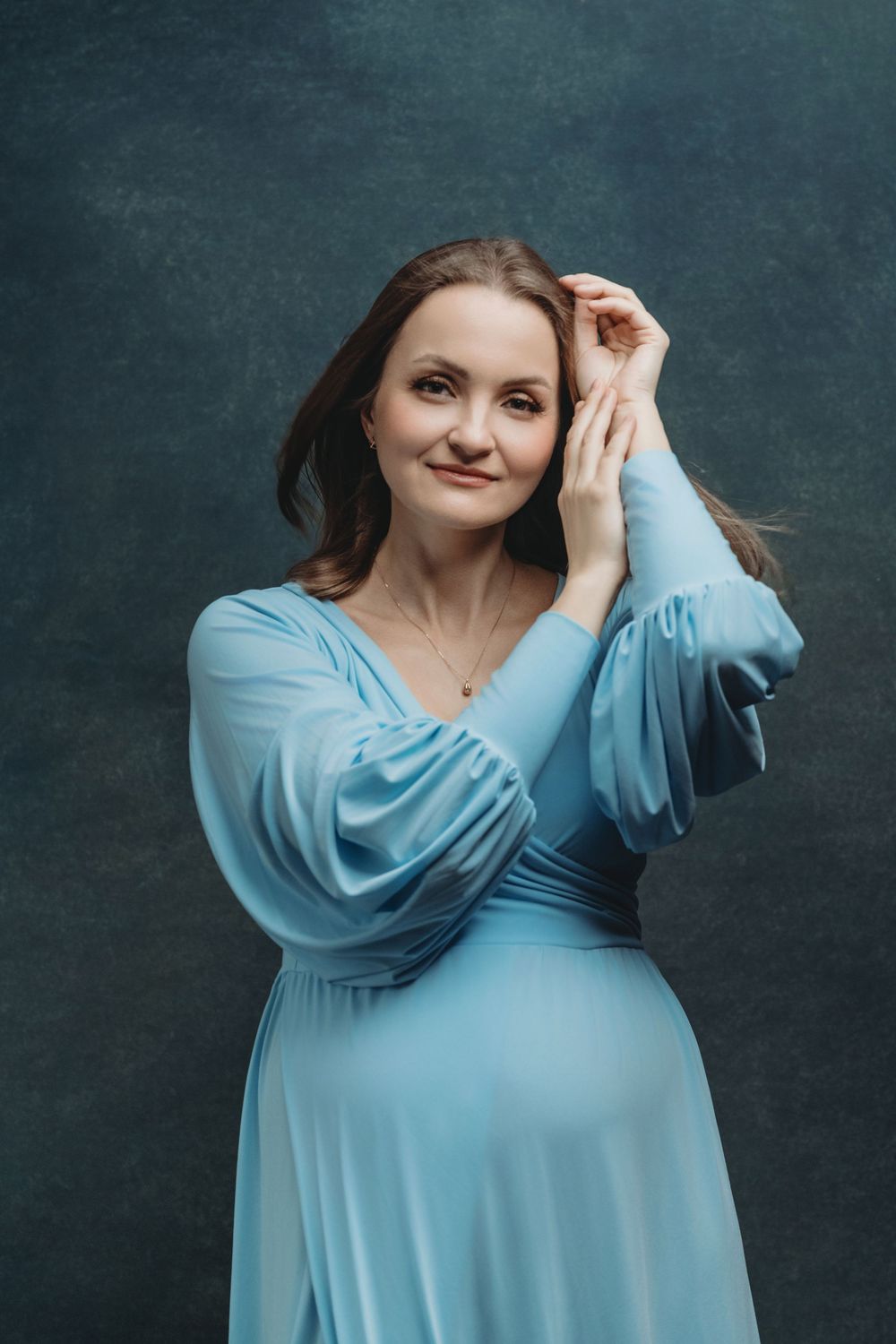 A maternity portrait of a woman in a flowing light blue dress posing against a dark backdrop - maternity photography Bellevue WA