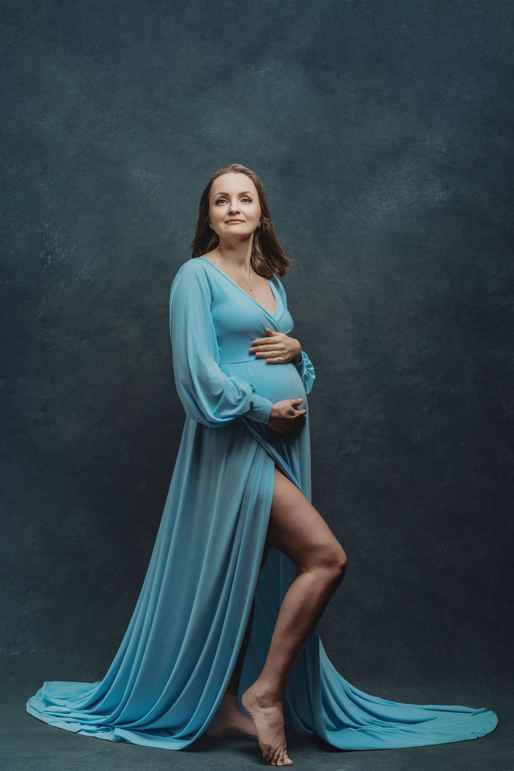 A flowing light blue maternity gown with sheer sleeves drapes elegantly against a dark moody backdrop - maternity photography Bellevue WA