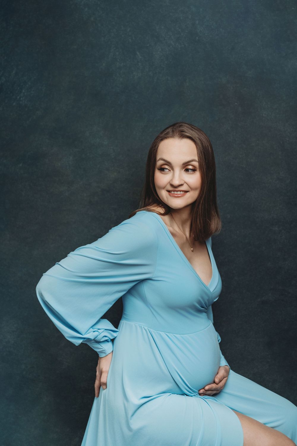 An expectant mother in a soft blue dress poses gracefully against a dark gray studio background - maternity photography Bellevue WA