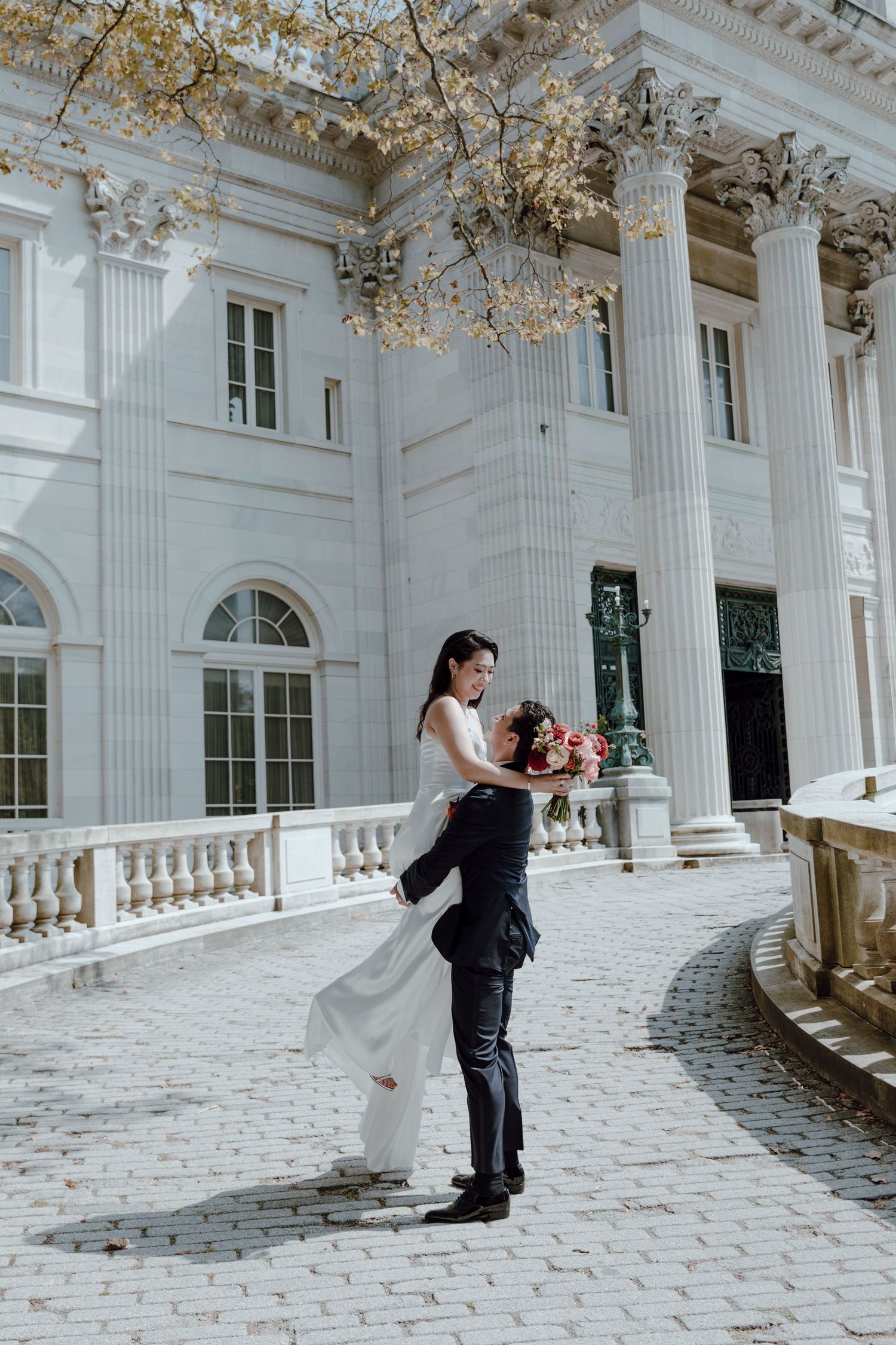 A couple shares a romantic dance moment in a grand white columned ballroom with ornate architectural details.