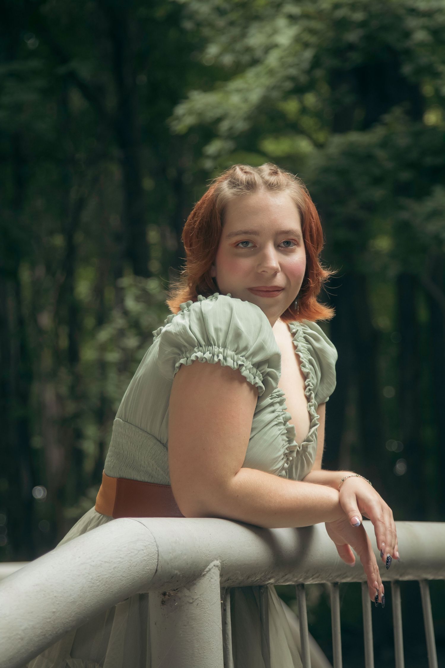 Woman in sage green ruffled top leans on railing in outdoor setting with forest background.