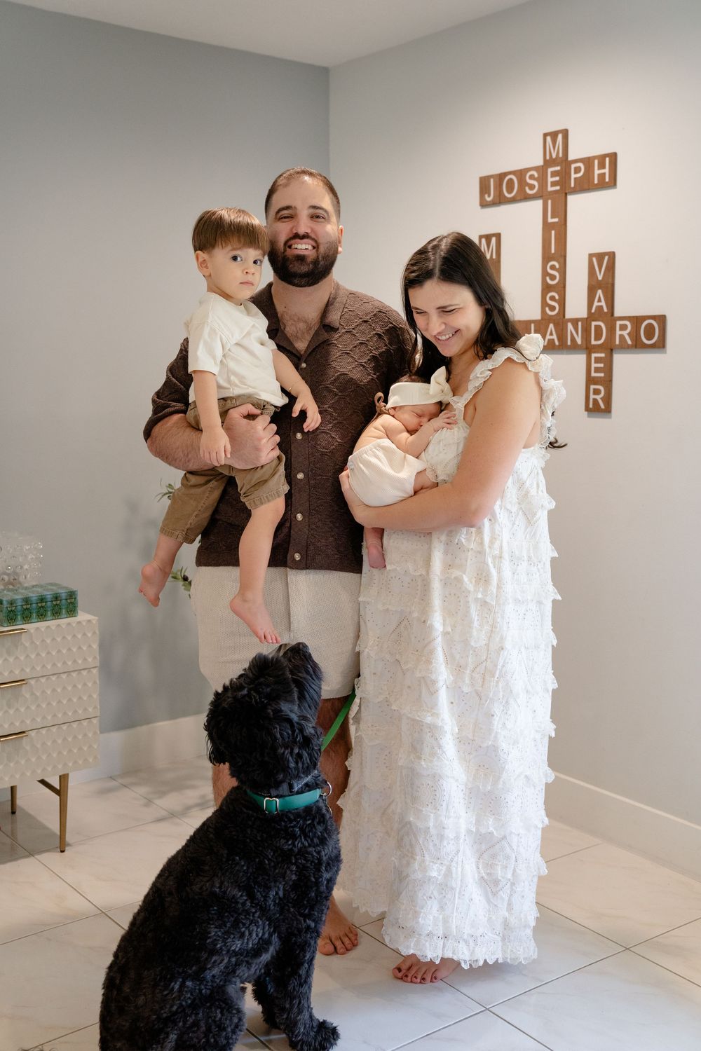 Family and black dog pose together in white nursery room with wooden cross decor on wall.