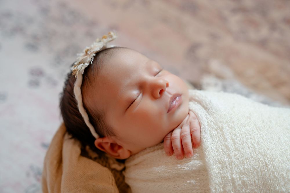 A sleeping newborn baby wrapped in white swaddling cloth lies peacefully on a textured beige surface.