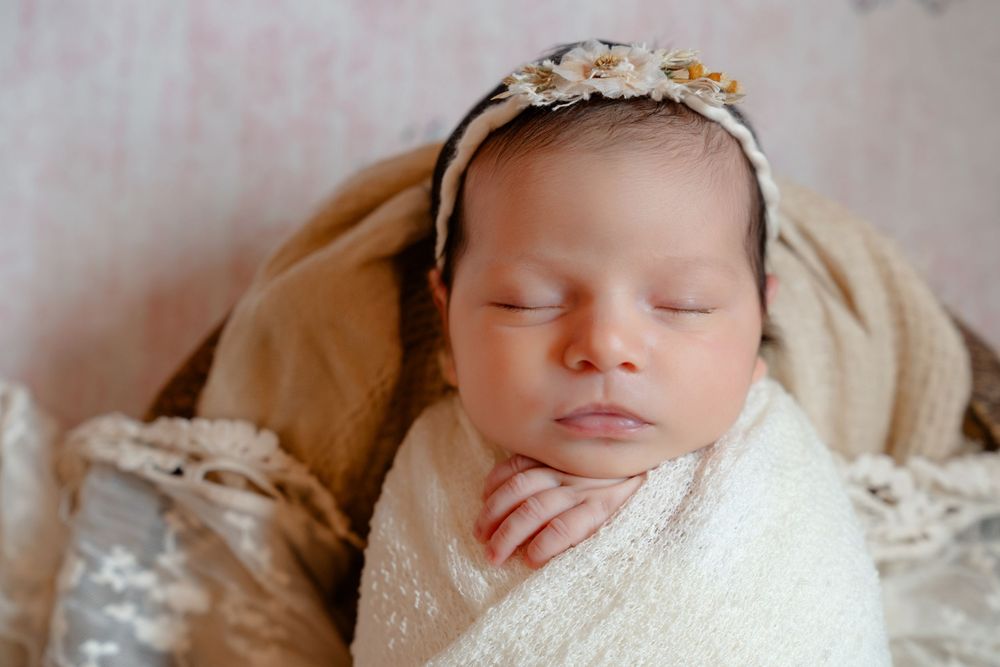 Sleeping newborn wrapped in white lace swaddle wearing delicate floral headband on beige backdrop.