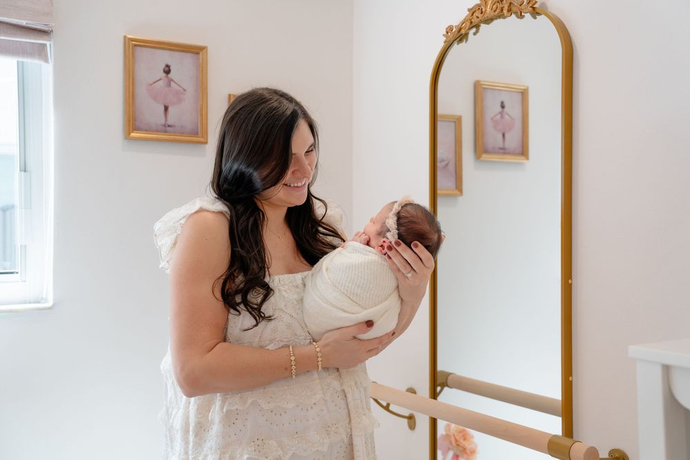 A woman in a white dress holds and gazes lovingly at her newborn baby in a bright nursery setting.