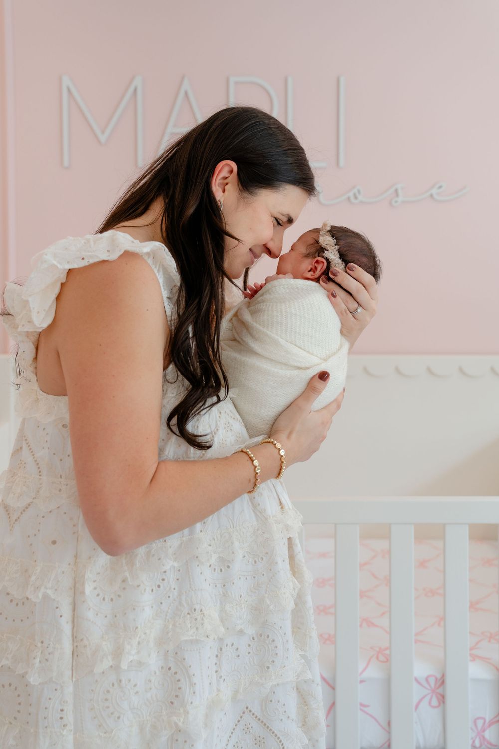 A tender moment between mother and baby in a soft pink nursery with white crib railing.