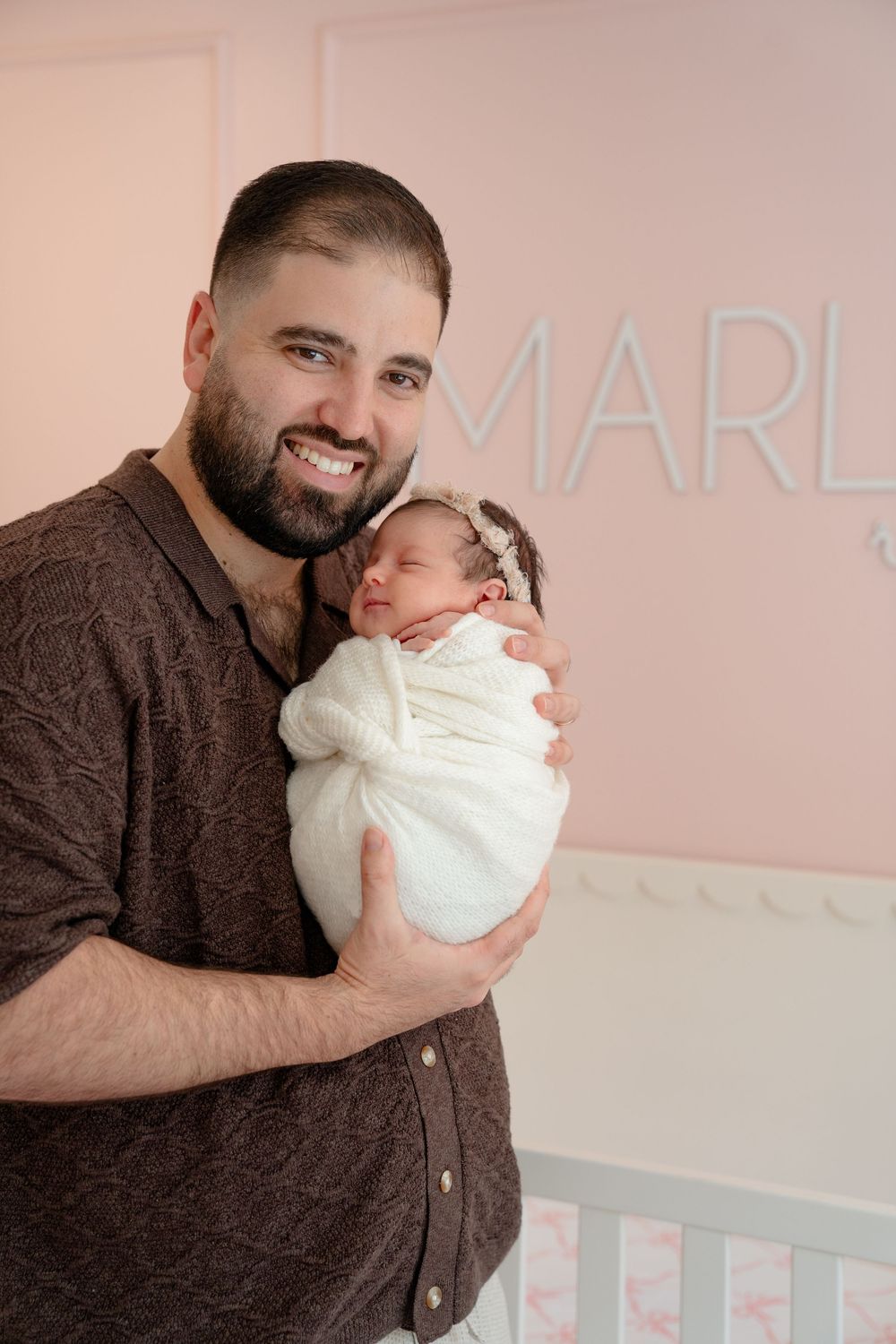 Parent in brown sweater cradling newborn baby wrapped in white blanket.