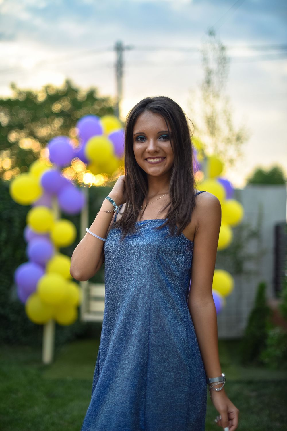 A person in a blue denim dress poses outdoors with yellow and purple balloons in the background during golden hour.