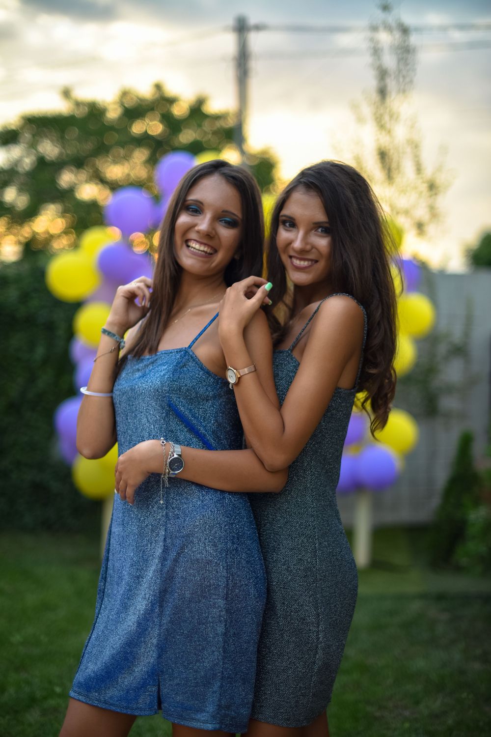 Two friends in blue dresses share laughter and hugs while posing outdoors with purple and yellow balloons at sunset.