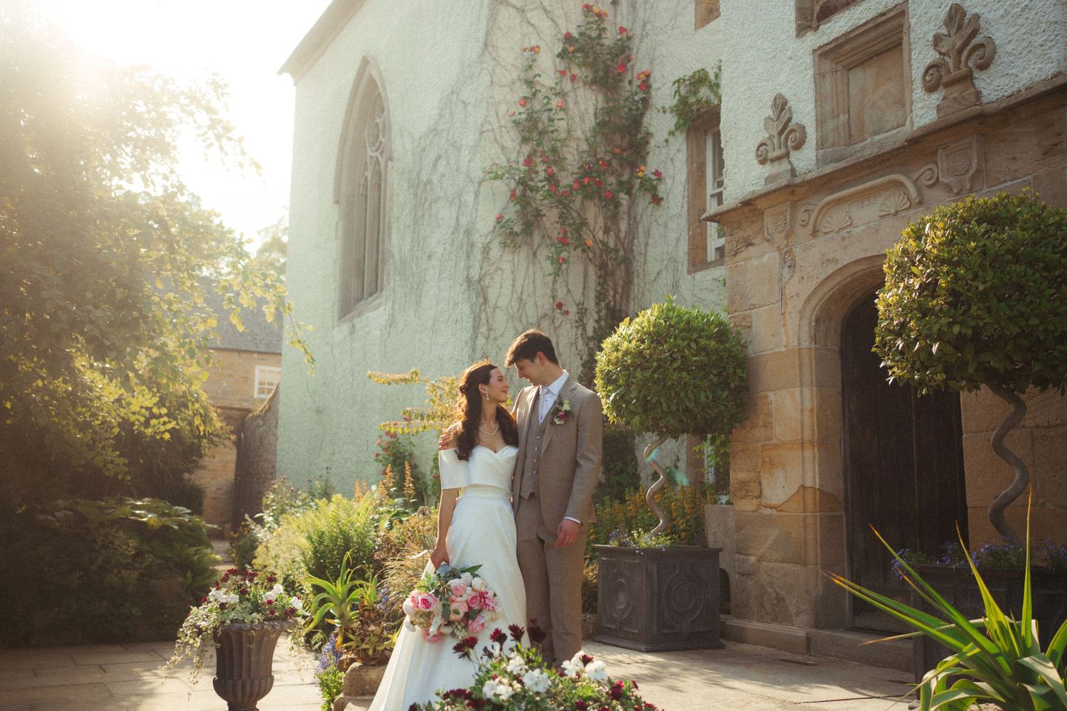 A couple shares a romantic moment in front of a stone church with lush gardens and golden sunlight.