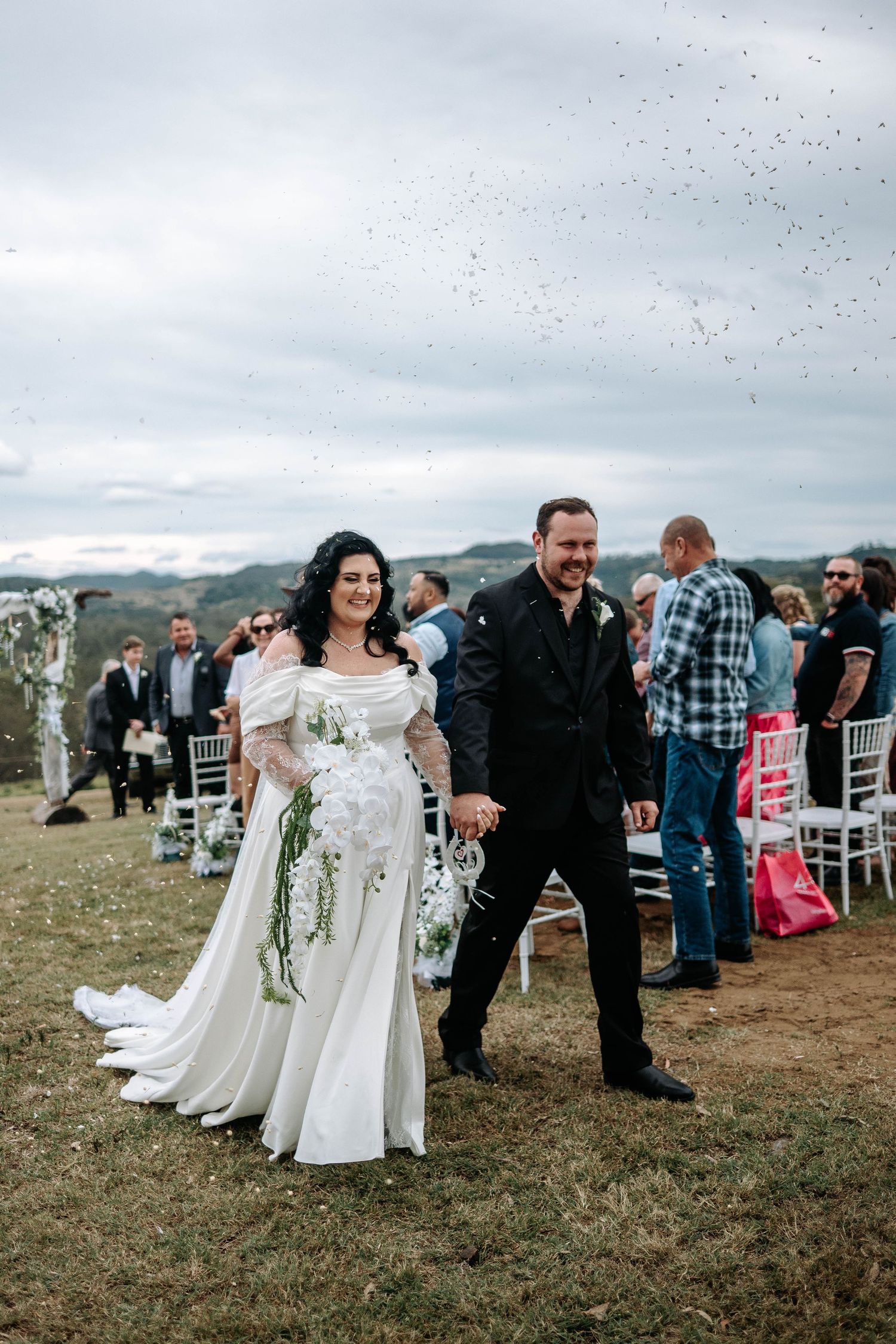 Wedding party gathers outdoors on grassy hill during cloudy day for group photos.