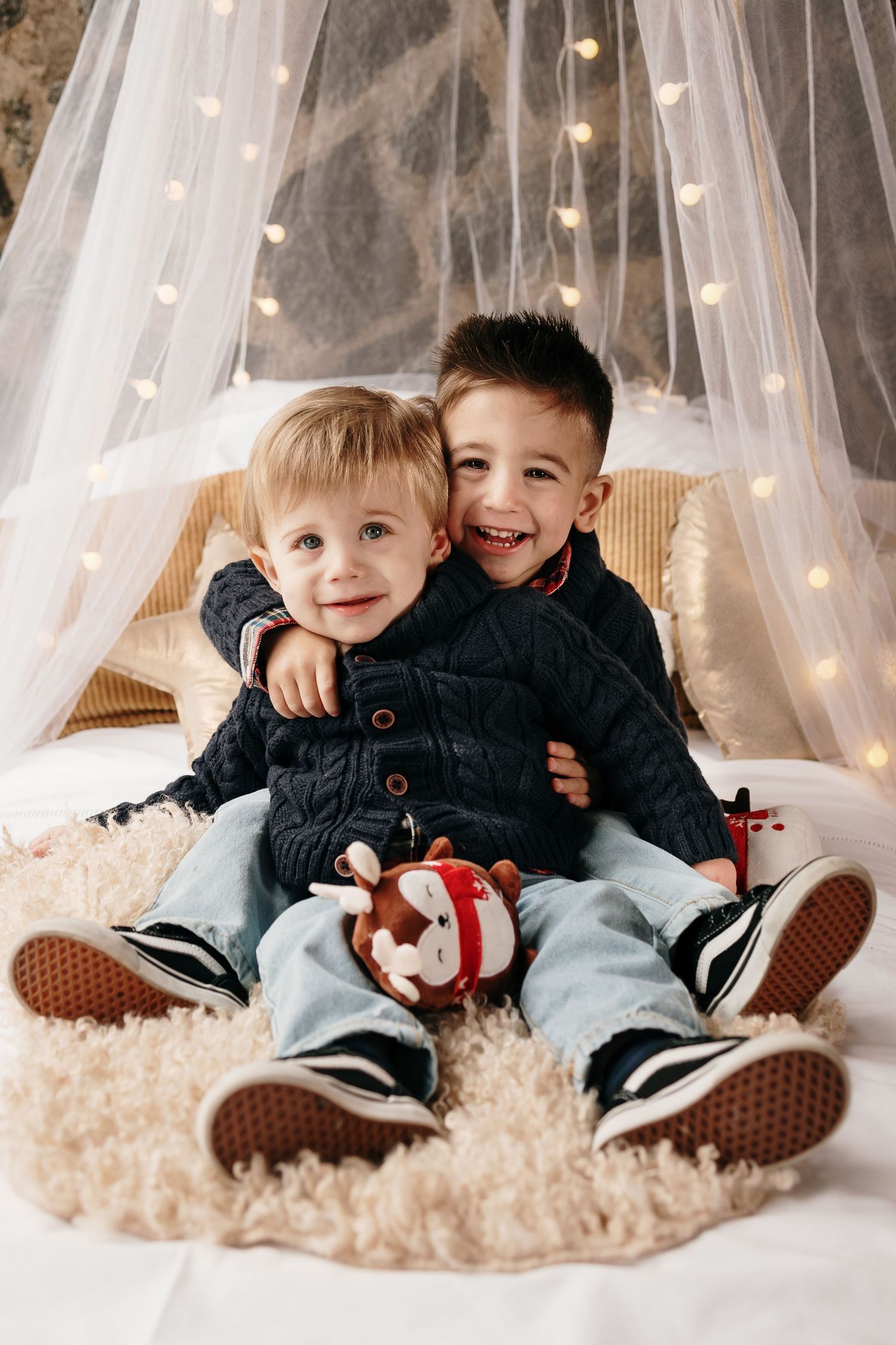 Two young children playing together on a fluffy white rug under string lights and sheer curtains.