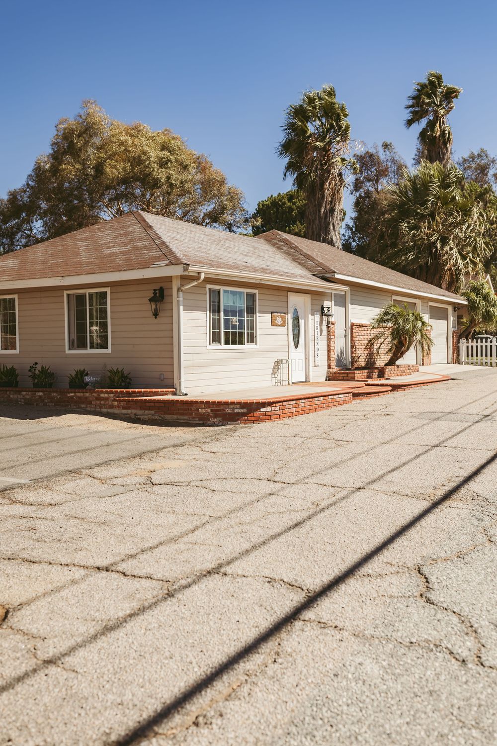 Modern single-story ranch house with tan siding and a large gravel driveway under blue sky with palm trees.