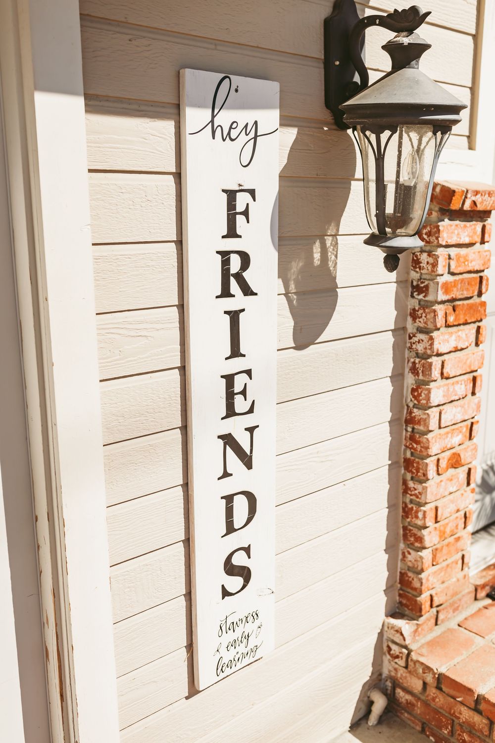 A vertical white wooden sign reading 'hey FRIENDS always stay humble' hangs beside a black outdoor lantern on a white wall.