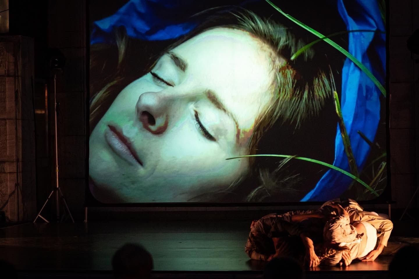 Close-up projection of sleeping woman on stage screen with performer lying below in theatrical production.
