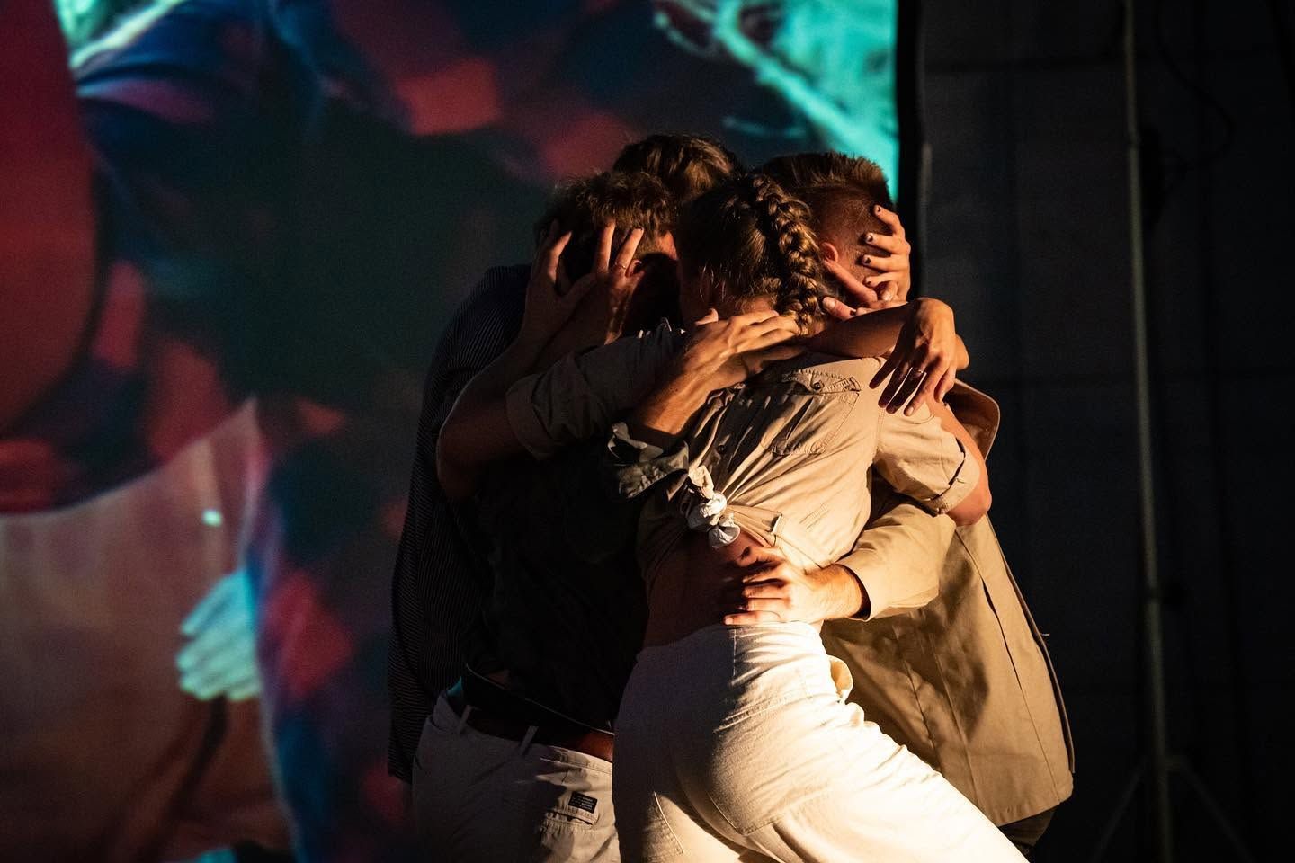 Three performers embracing in emotional group hug on stage with colorful backdrop during live performance.