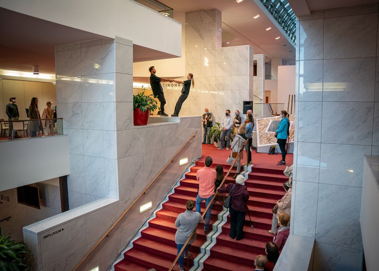 Performers on grand staircase with red carpet in elegant marble theater building interior.
