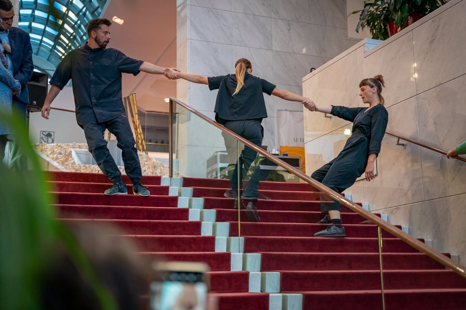 Three dancers holding hands descending red carpeted staircase in grand marble theater lobby.