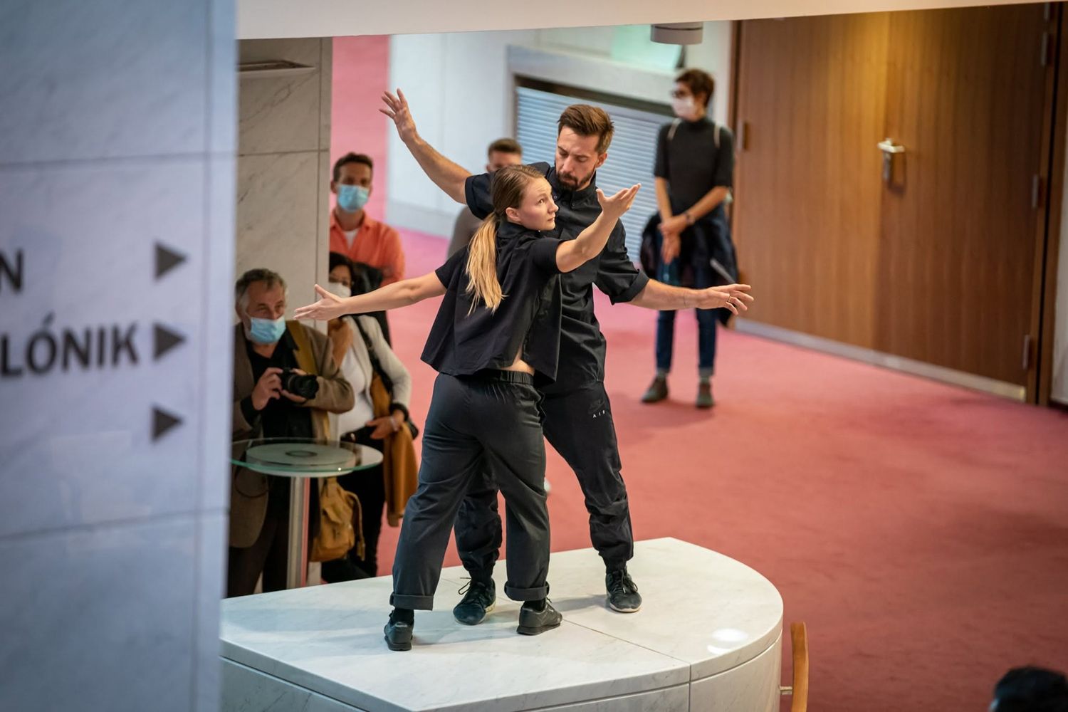 Two dancers in black performing contemporary dance on circular platform in modern theater lobby.