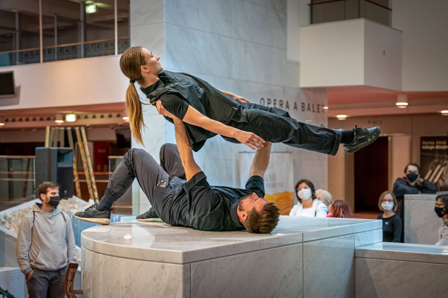Male dancer balancing female performer horizontally in acrobatic lift on marble surface in theater.