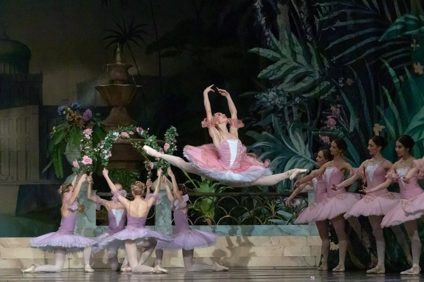 Ballet dancer leaping gracefully in pink tutu surrounded by ensemble in classical ballet performance.