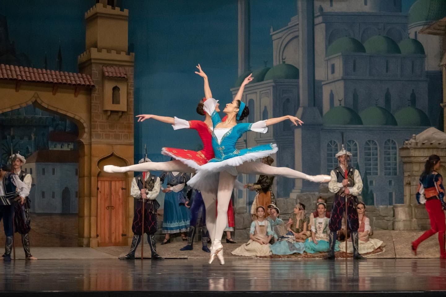 Two ballet dancers performing grand jete in colorful exotic stage set with full cast ensemble.