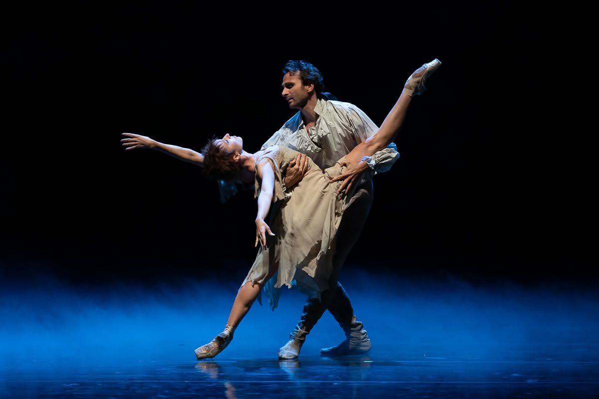 Two contemporary dancers perform dramatic lift on dimly lit stage, woman arching back gracefully in flowing costume.
