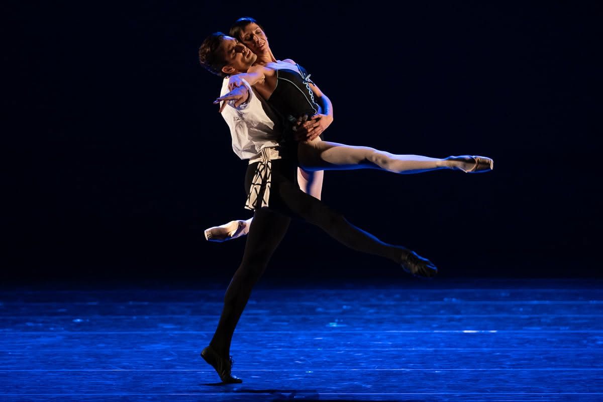 Two ballet dancers performing a dramatic lift on a blue-lit stage in elegant costumes.
