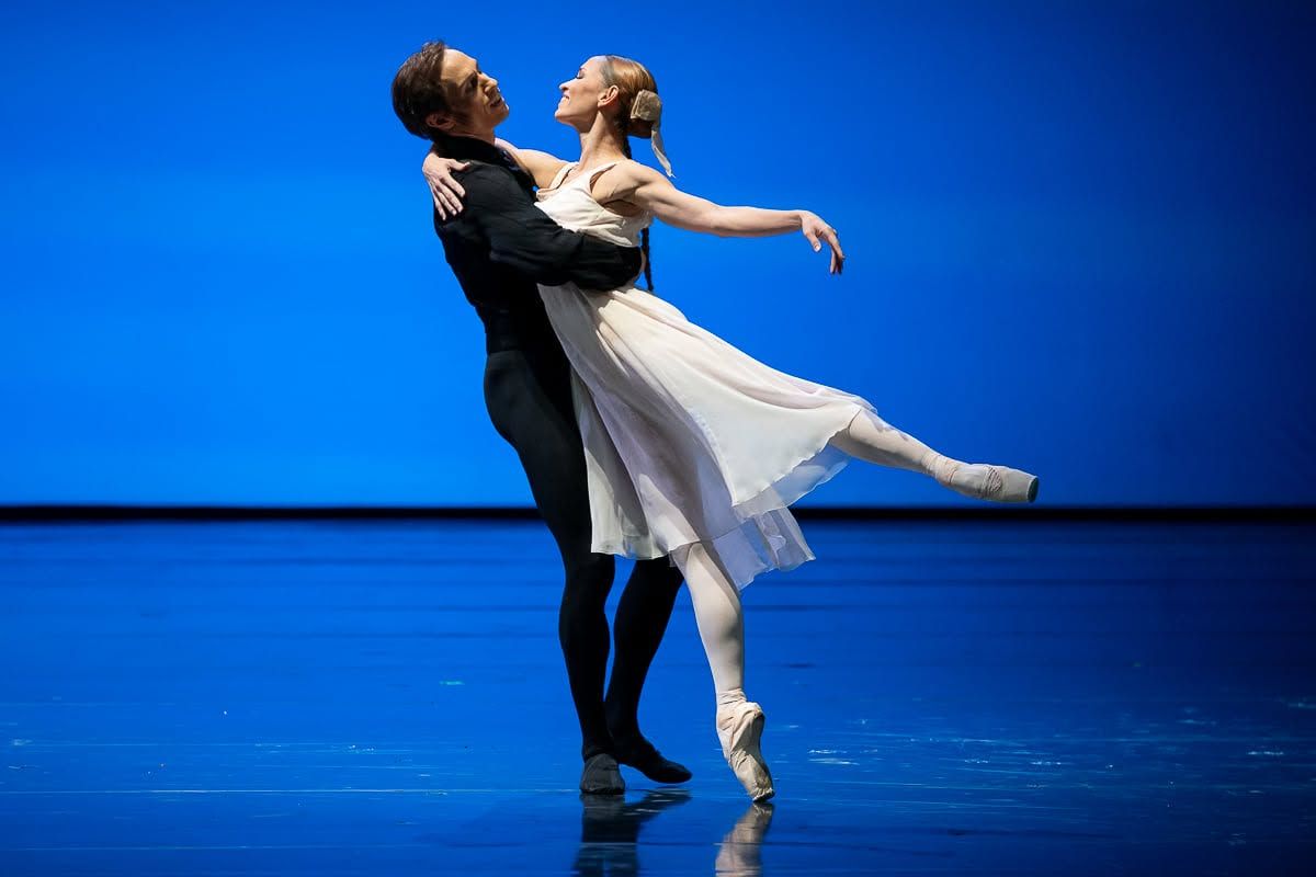 Ballet couple performing an elegant embrace on pointe on a vivid blue-lit stage.