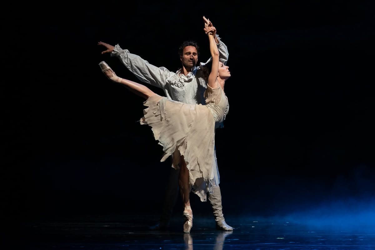 Male ballet dancer lifts female partner in tattered white tutu during dramatic performance on dark stage.