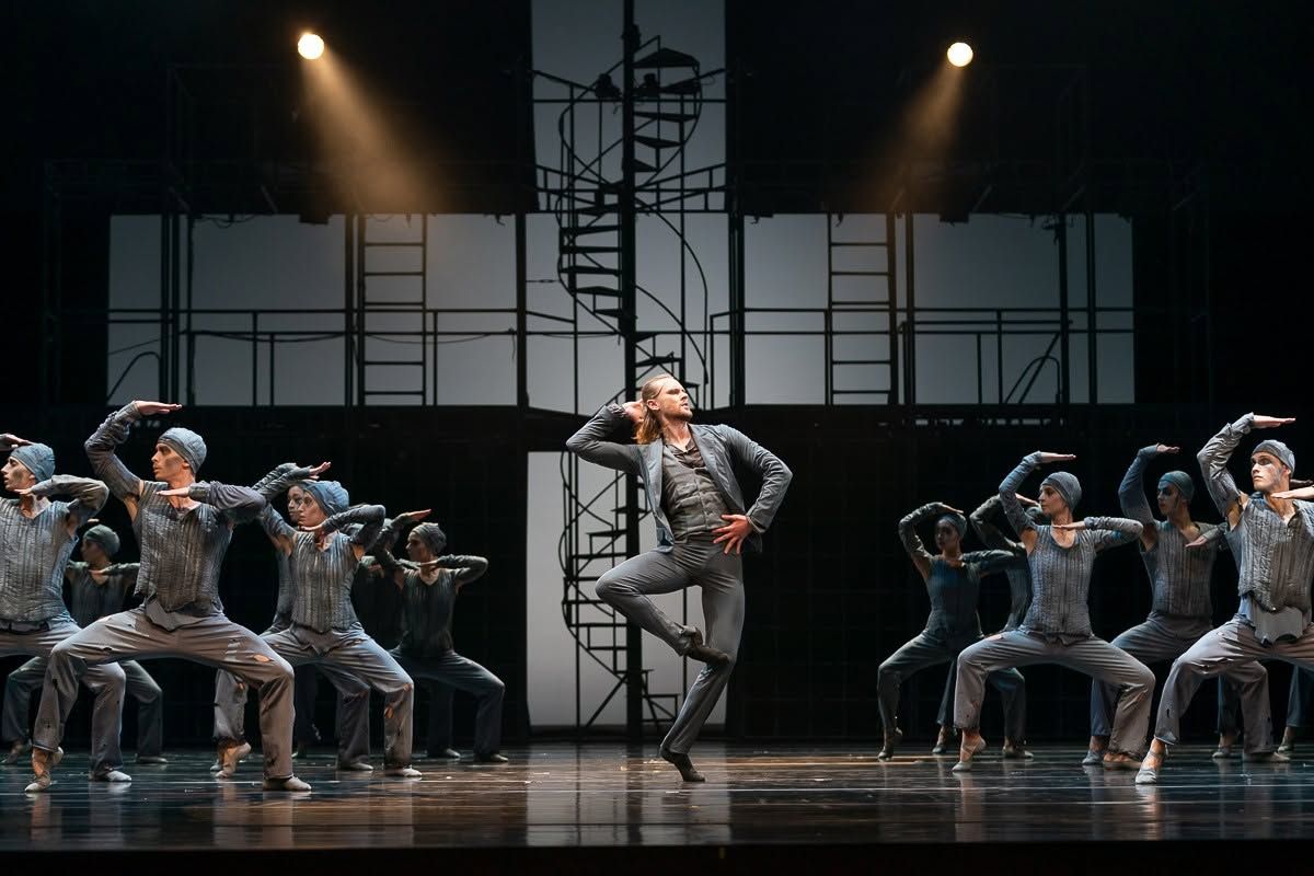 Lead dancer performs center stage surrounded by ensemble in grey costumes beside industrial spiral staircase set.