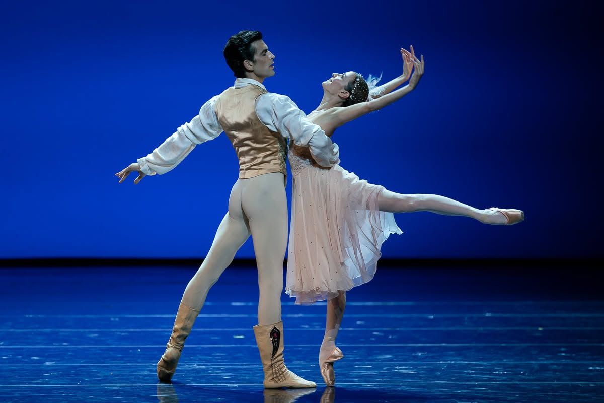 Elegant ballet pas de deux with male dancer supporting ballerina in flowing pink dress against bright blue backdrop.