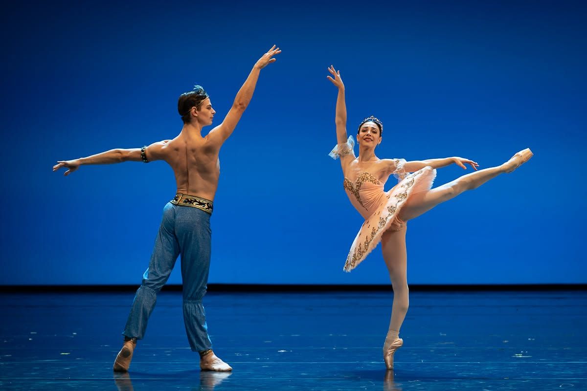 Two ballet dancers perform on stage, woman in pink tutu en pointe, male partner in jeans raises arm against blue backdrop.