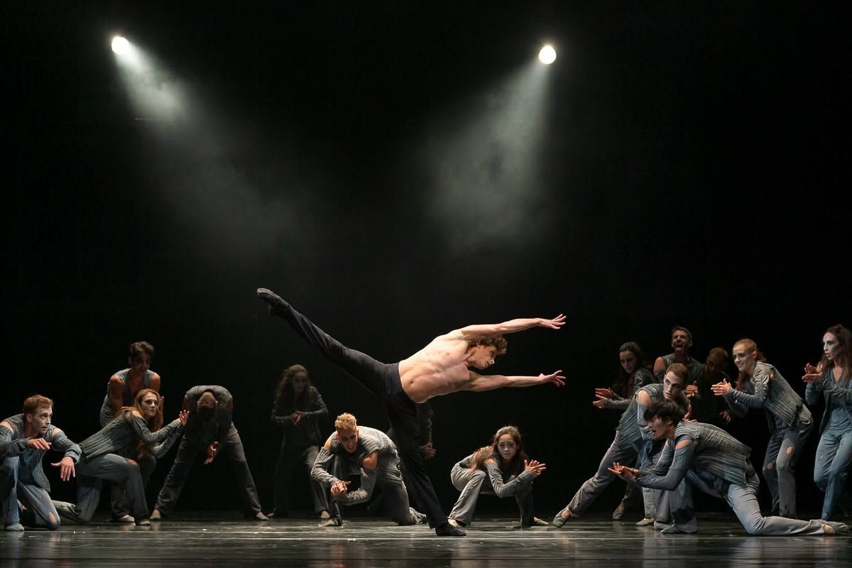 Shirtless male dancer leaps horizontally above crouching ensemble cast under dramatic spotlights on dark stage.