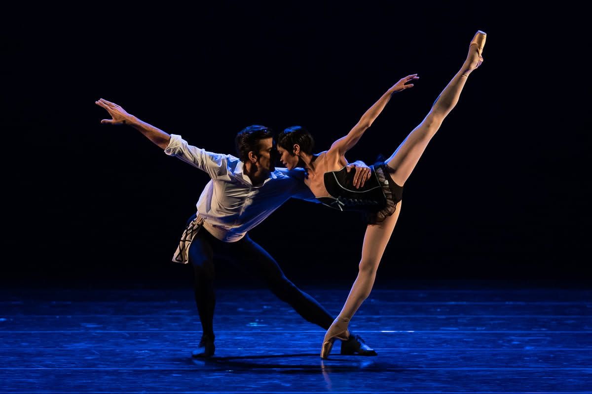Ballet couple performing an expressive contemporary lift on a dramatic blue-lit stage.