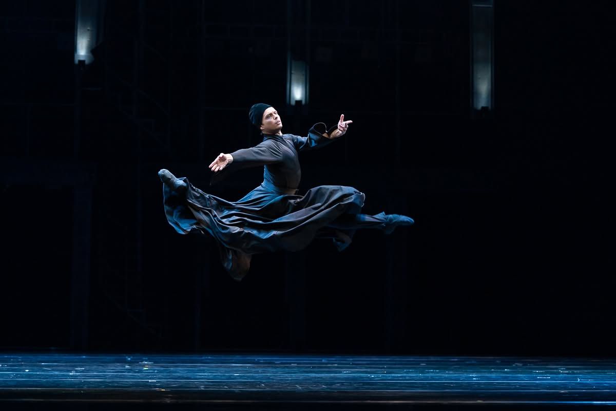 Solo dancer leaps dramatically in dark blue costume against black stage backdrop under dim lighting.