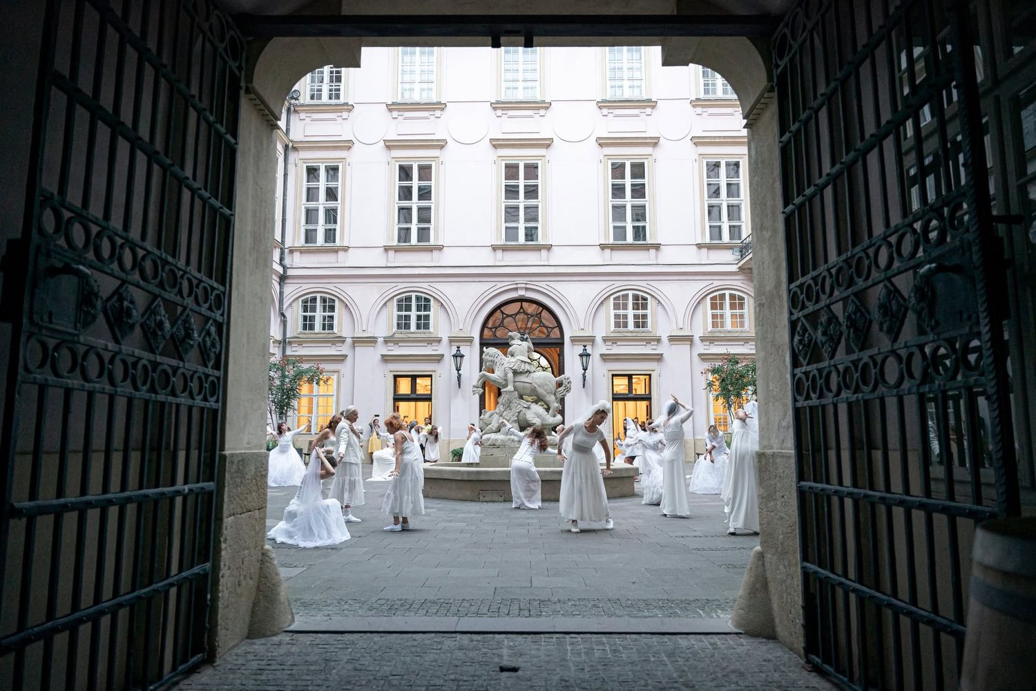 Performers in white costumes gather in a grand European courtyard during an elegant outdoor classical music event.