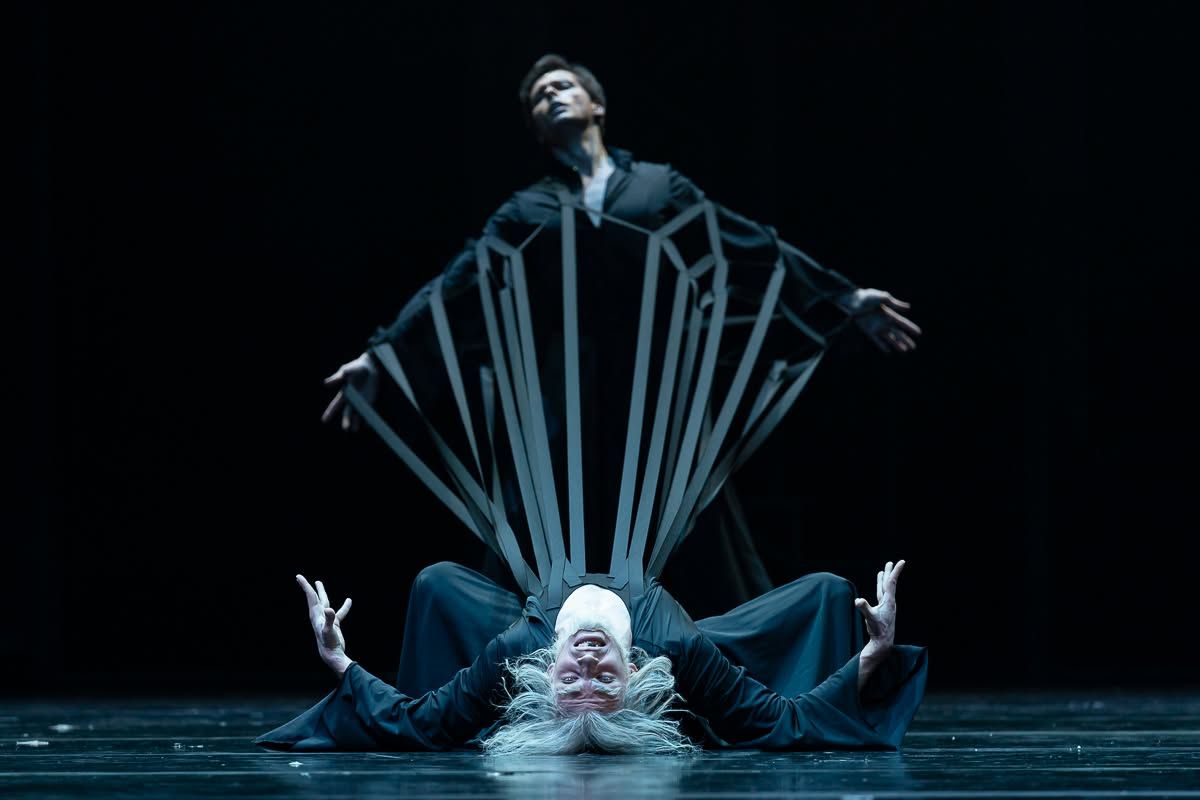 Two dancers in striking black geometric costumes perform dramatic floor and standing poses on dark stage.