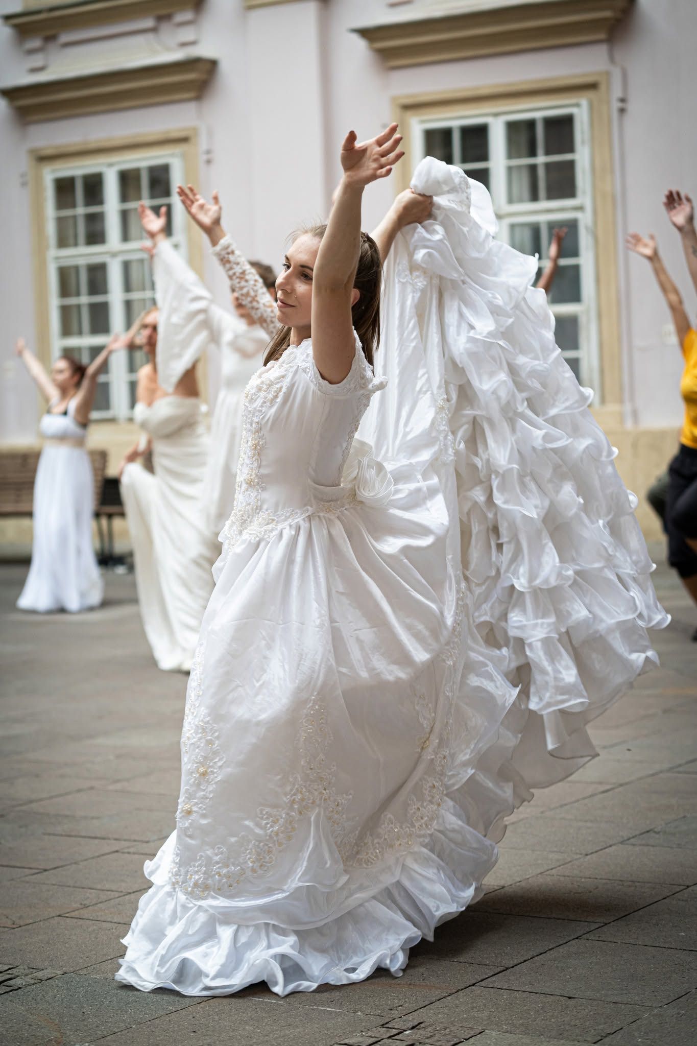 A woman in a flowing white wedding dress dances with arms raised outdoors at a courtyard.