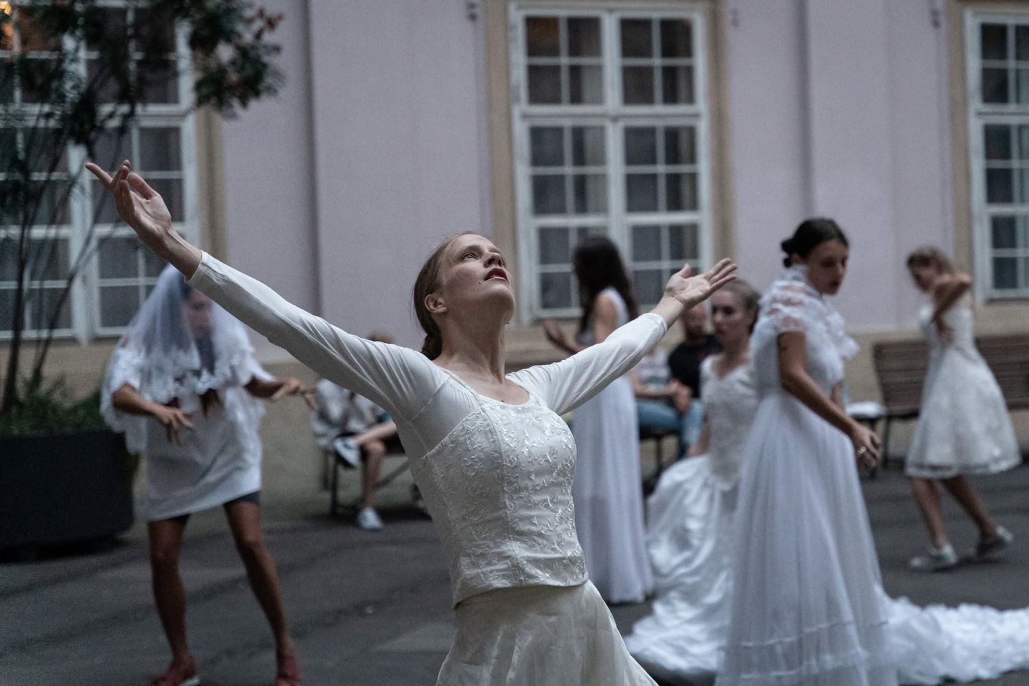 A woman in a white wedding dress dances with arms outstretched in an outdoor courtyard performance.