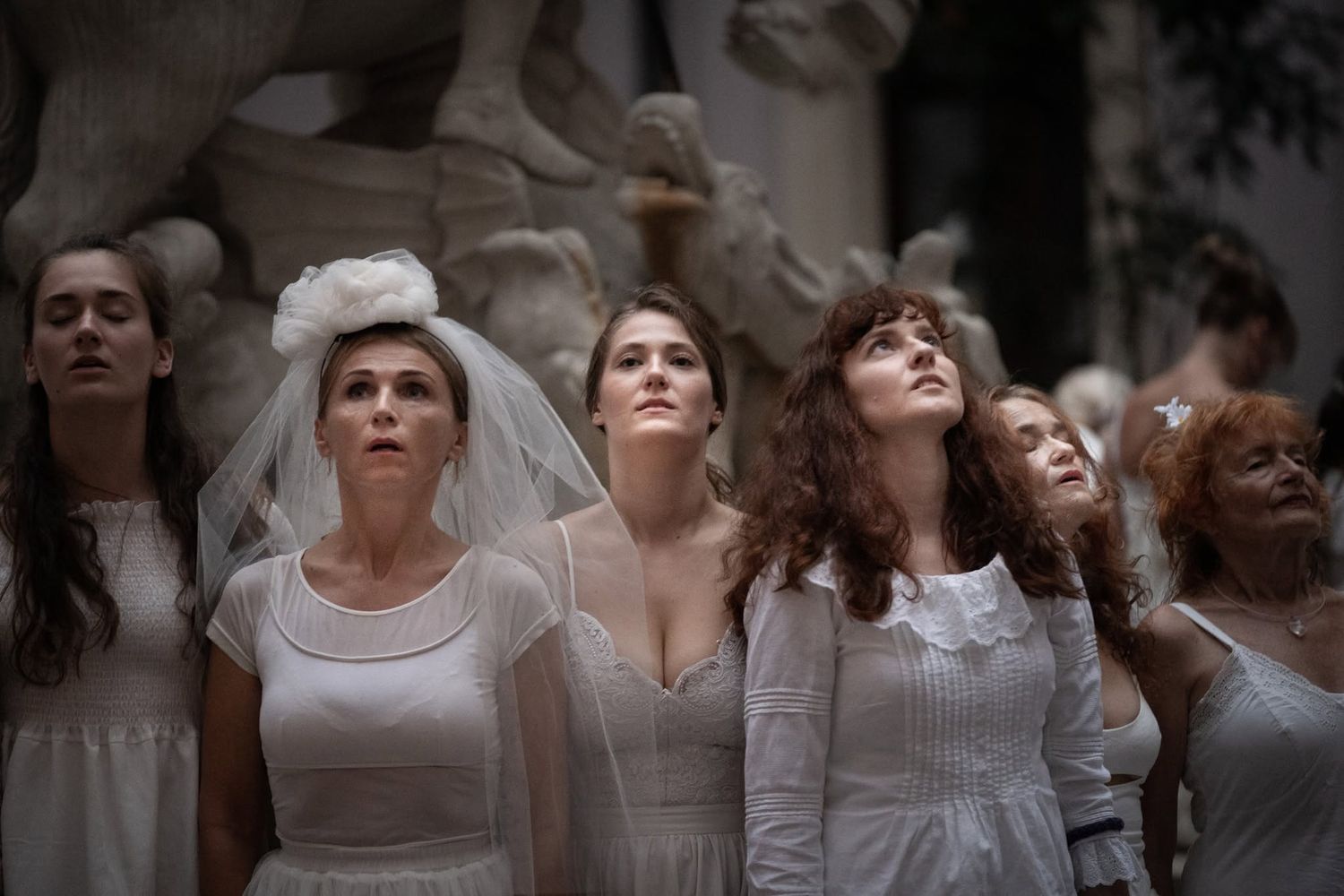 A group of women dressed in white bridal gowns and veils pose solemnly outdoors.