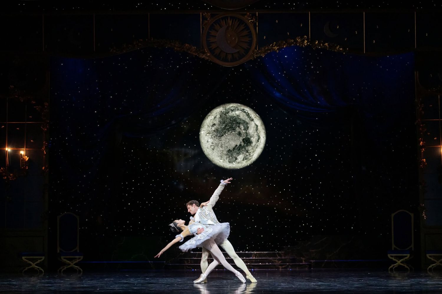 Ballet pas de deux performed before large illuminated moon backdrop in classical theatrical stage setting.