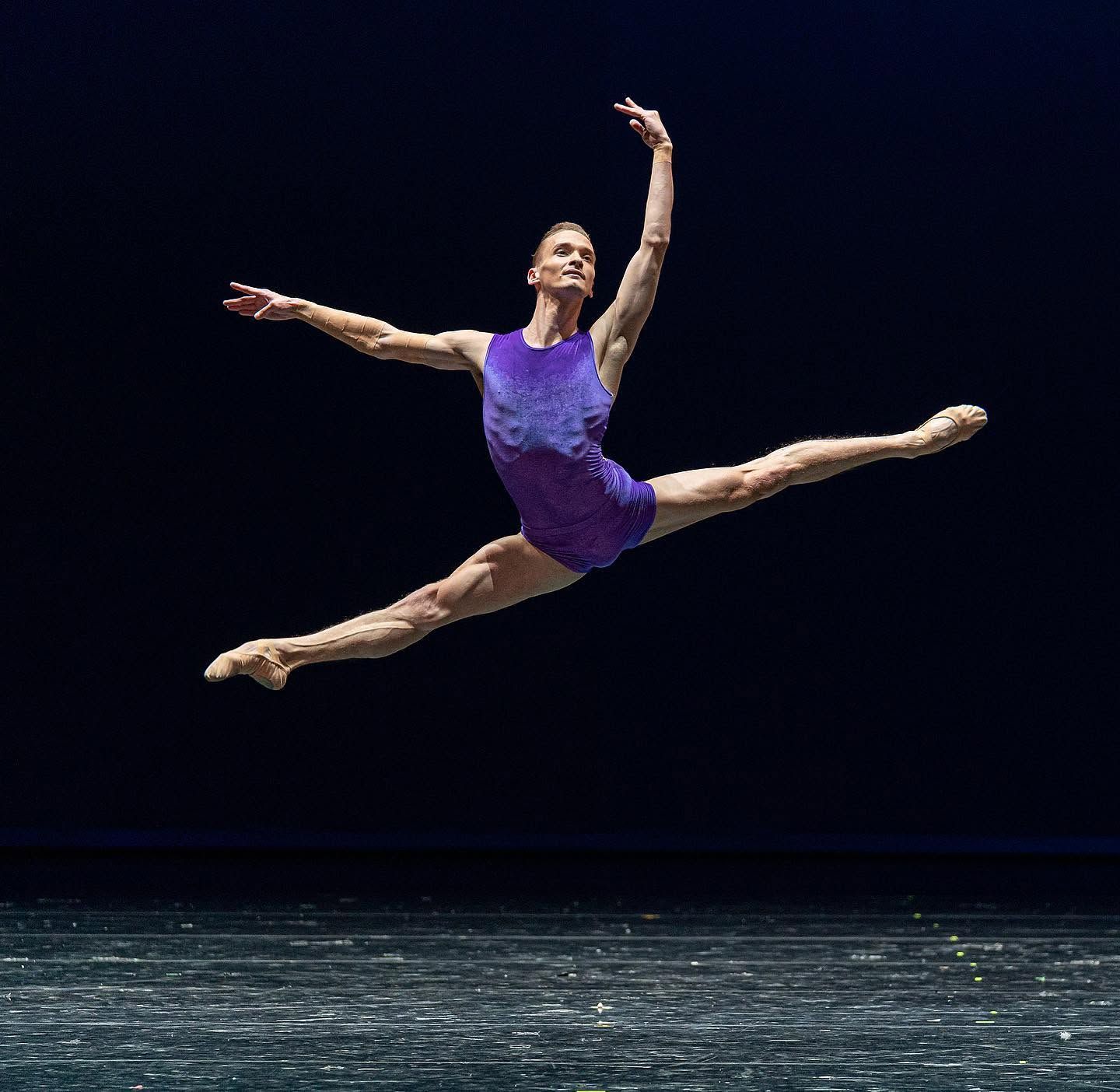 A male ballet dancer performs a grand jeté leap in a purple leotard on a professional stage.