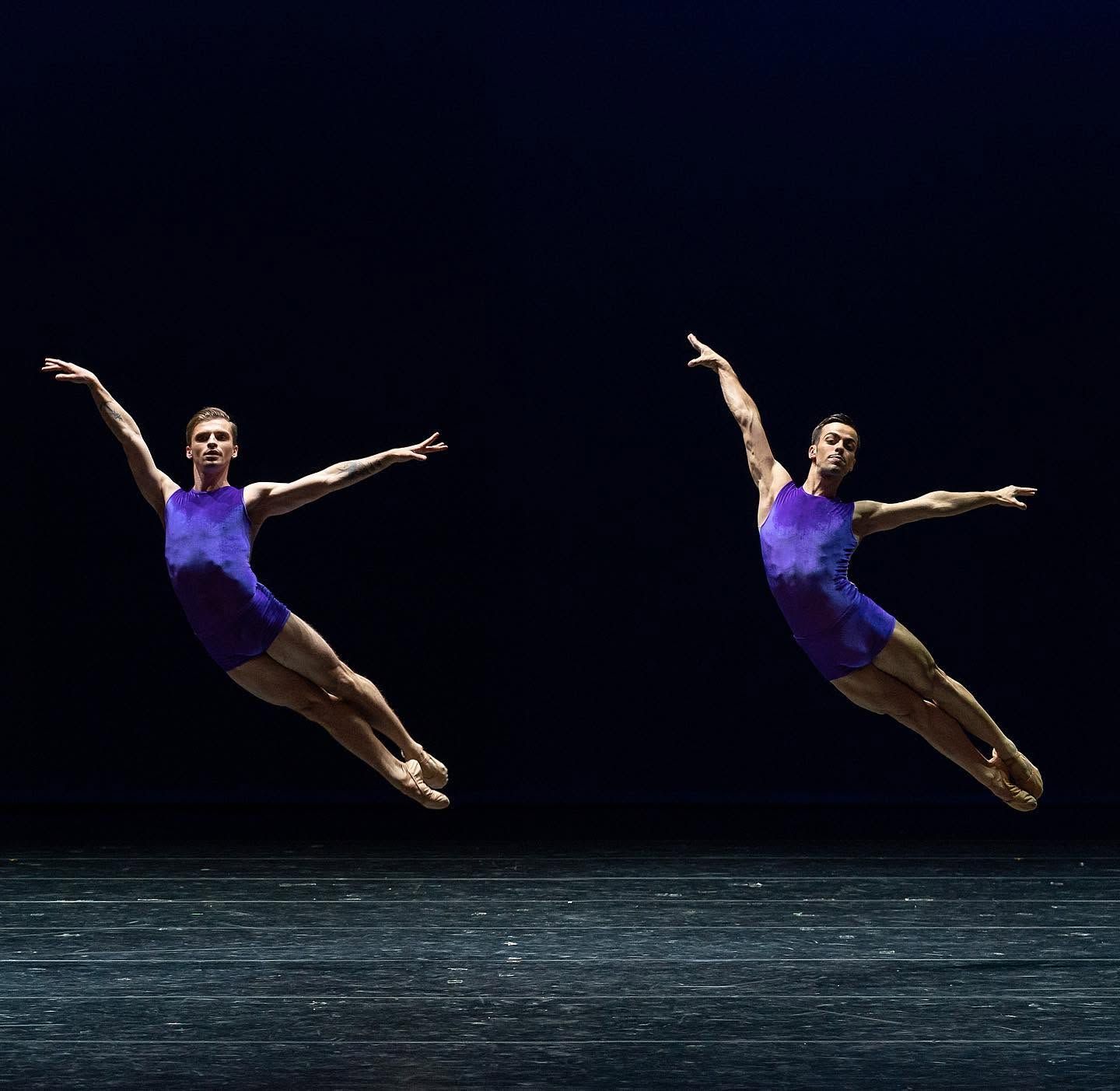 Two male ballet dancers leap gracefully in purple leotards against a dark stage background.