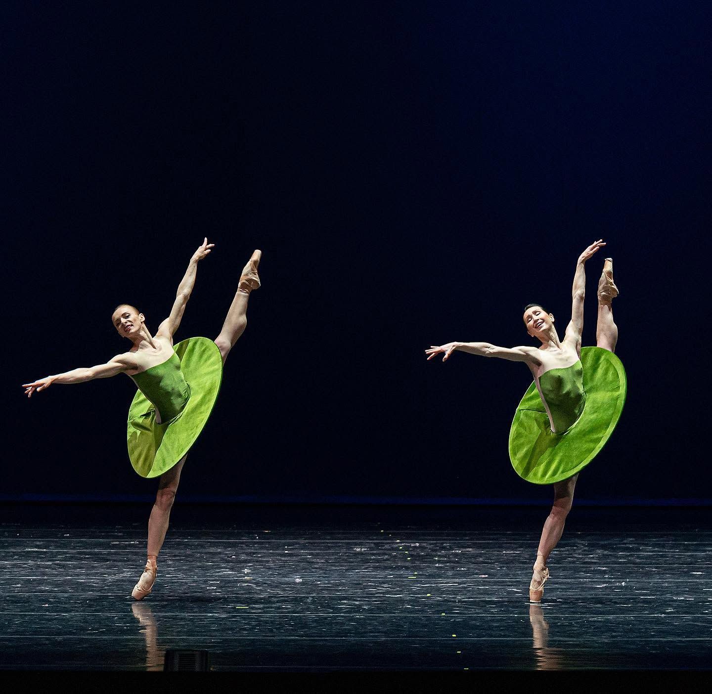 Two female ballet dancers perform an arabesque in matching green tutus on a dark stage.