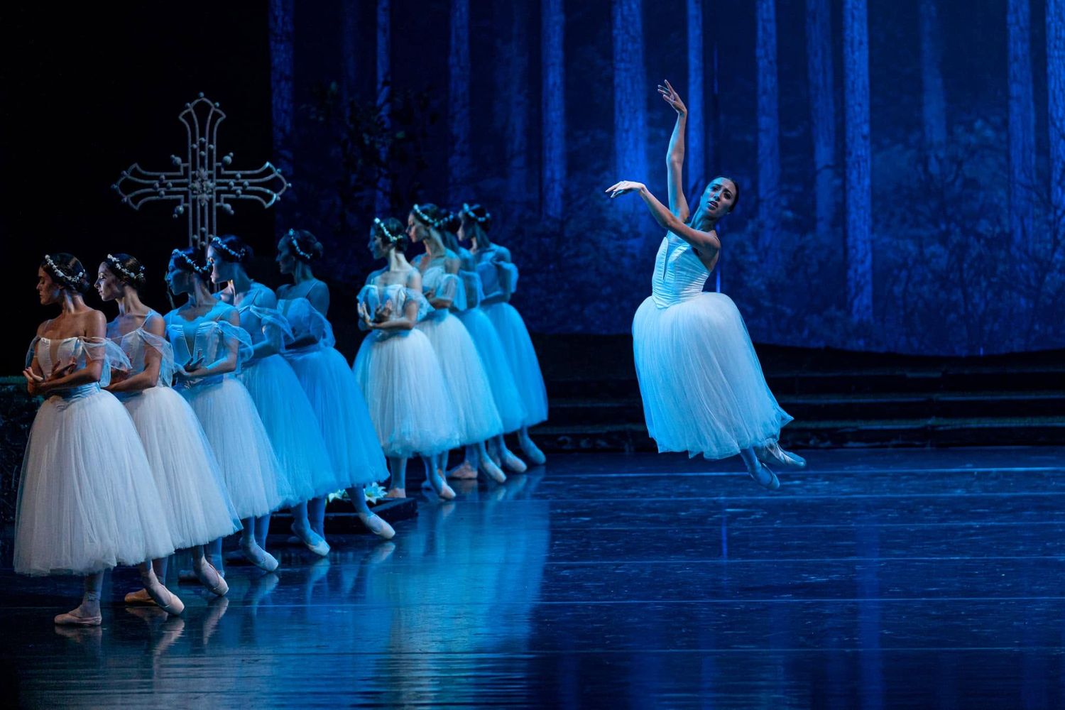 Ballet dancers in white tutus perform Giselle on a blue-lit stage with a cross prop in the background.