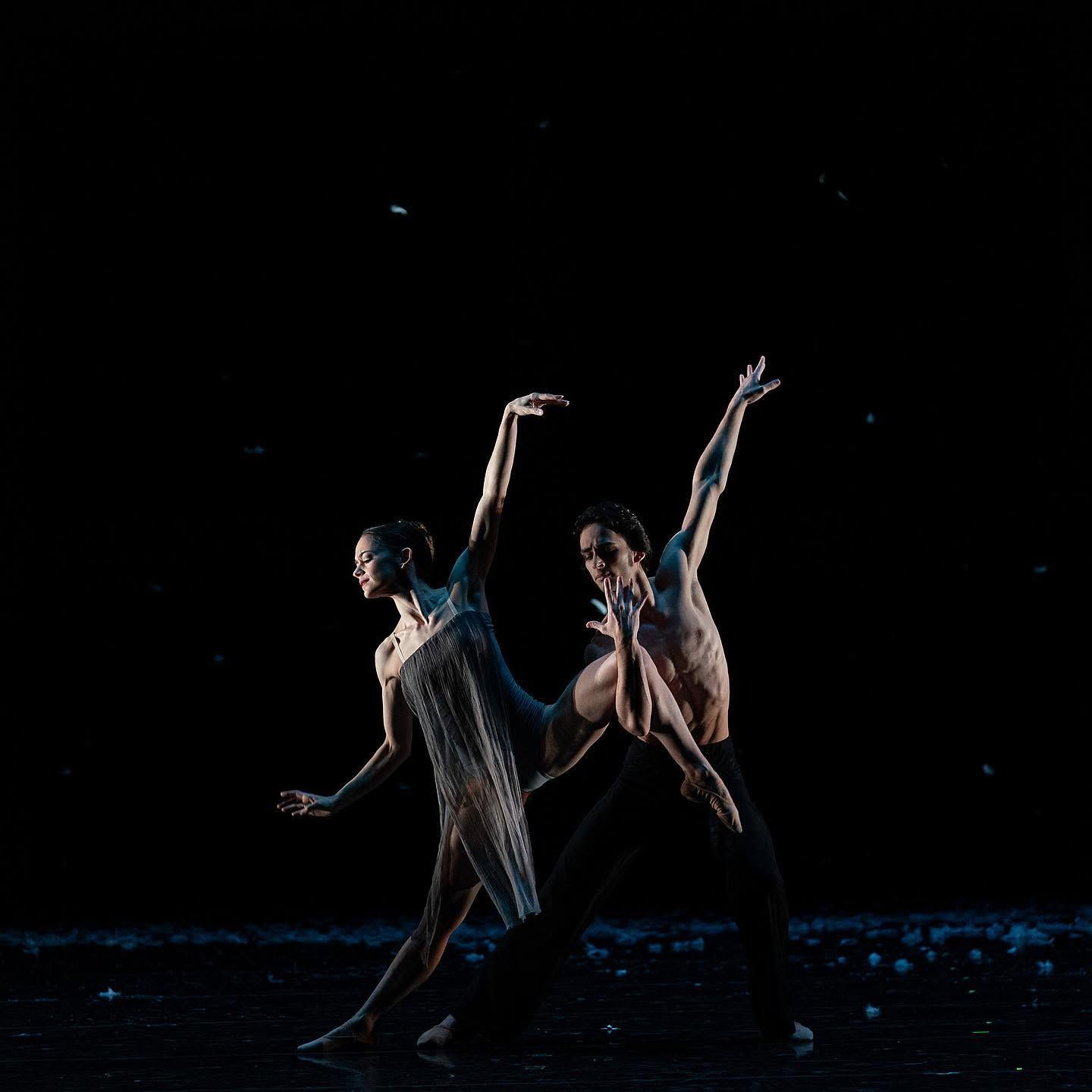 Two contemporary dancers extend arms gracefully in mirrored poses against black background with scattered light.