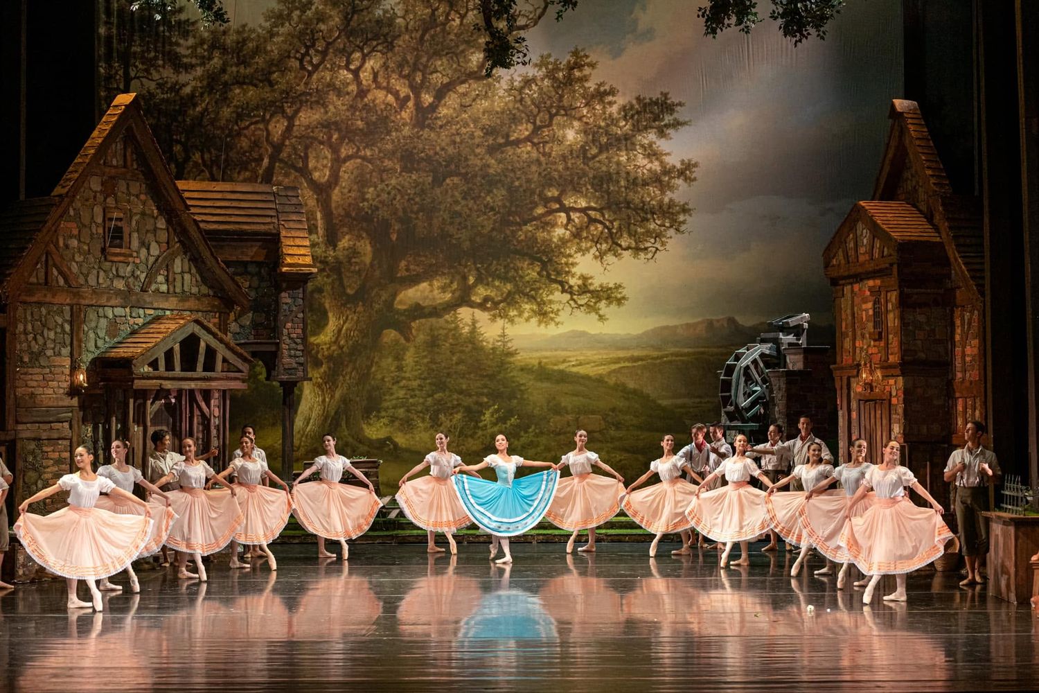 Ballet ensemble performs in colorful costumes before a scenic village backdrop with a large oak tree on stage.