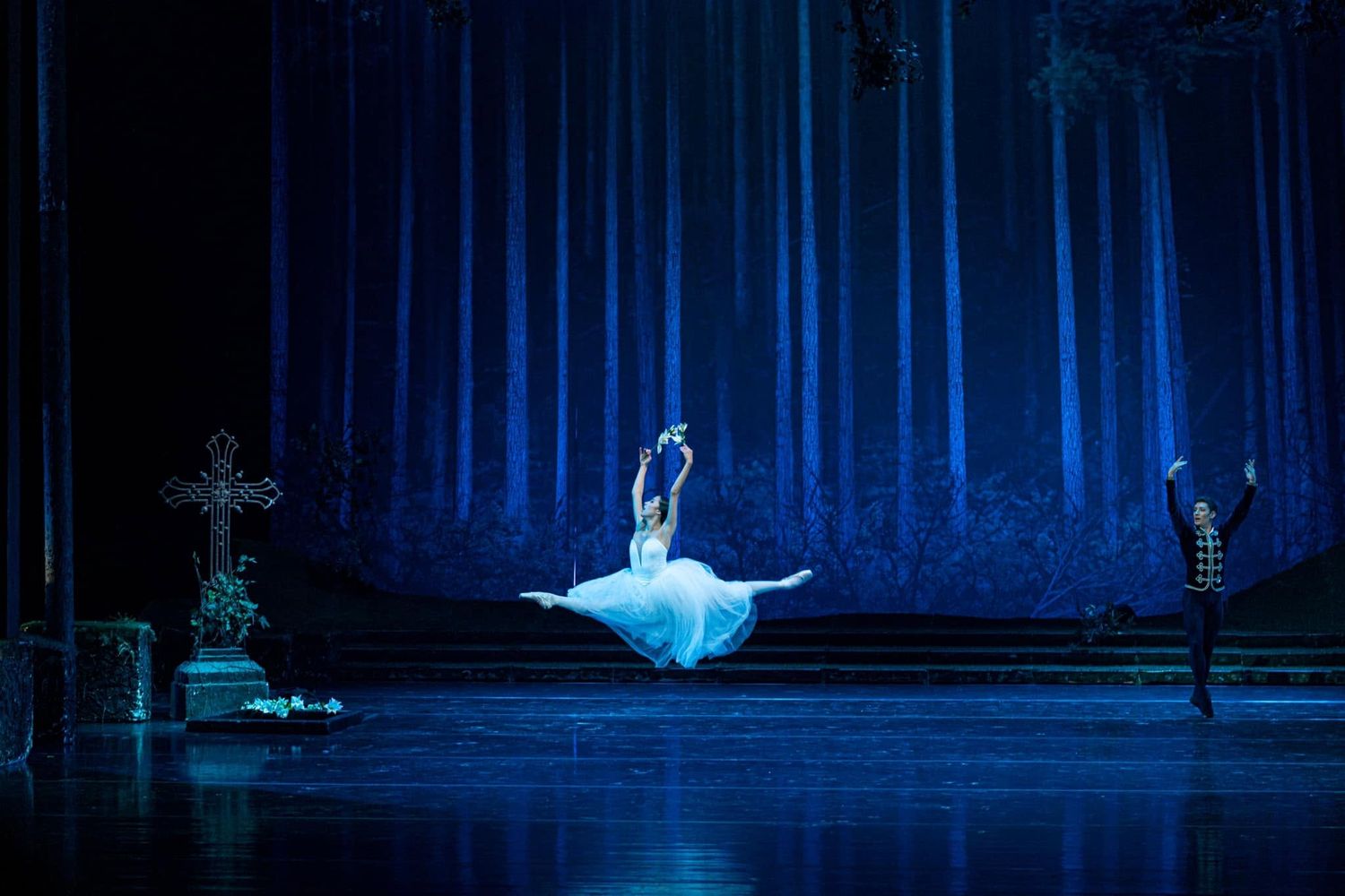 A ballerina leaps gracefully in a white tutu on a dramatic blue-lit stage during a classical ballet performance.