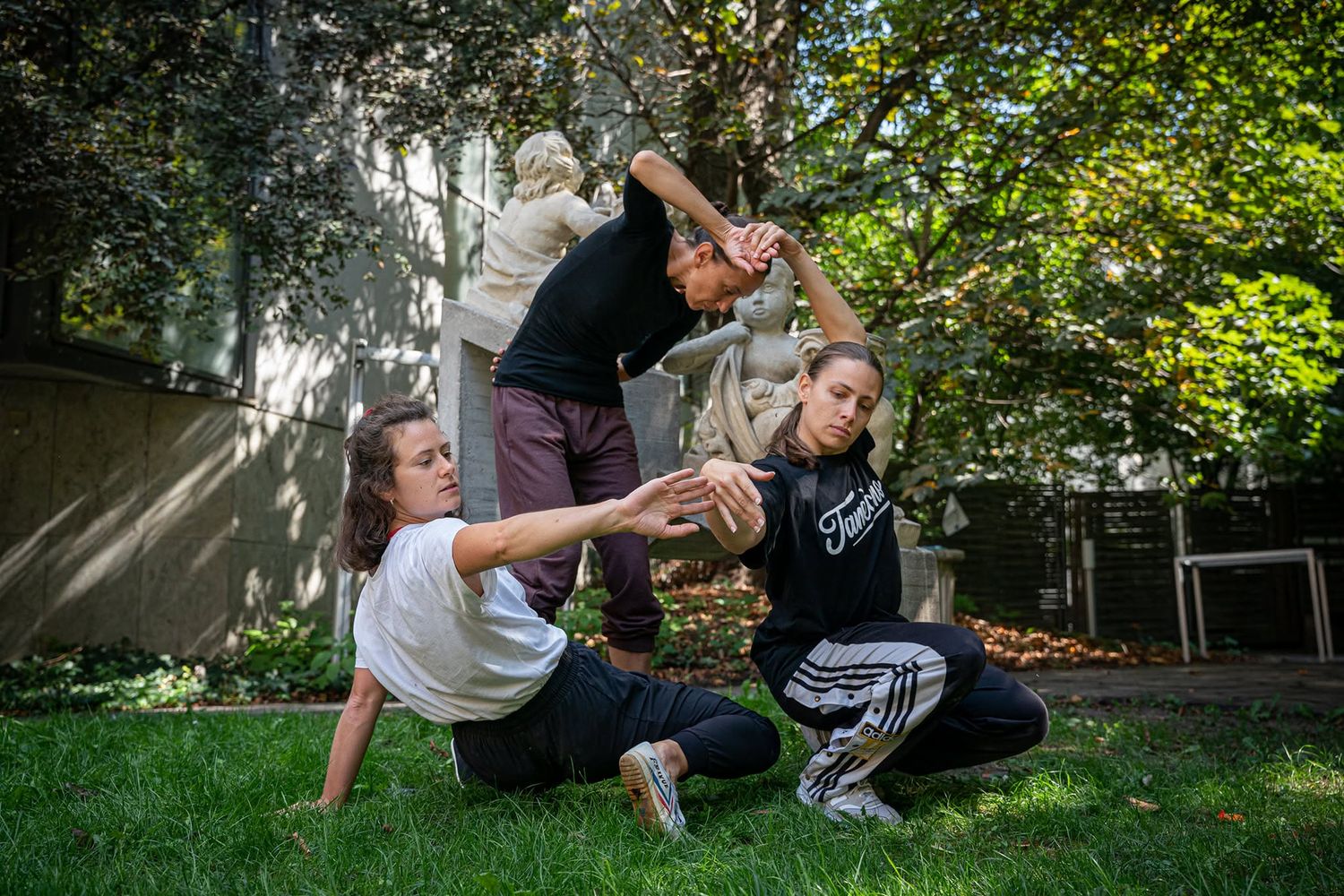 Three dancers rehearsing contemporary dance outdoors in a lush green garden setting.